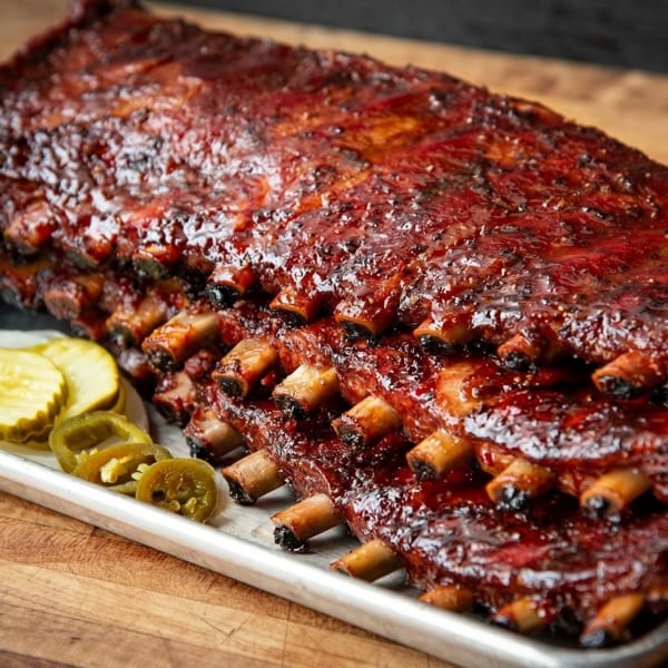 Rack of BBQ ribs with glistening sauce, served with sliced pickles and jalapeños on a metal tray.